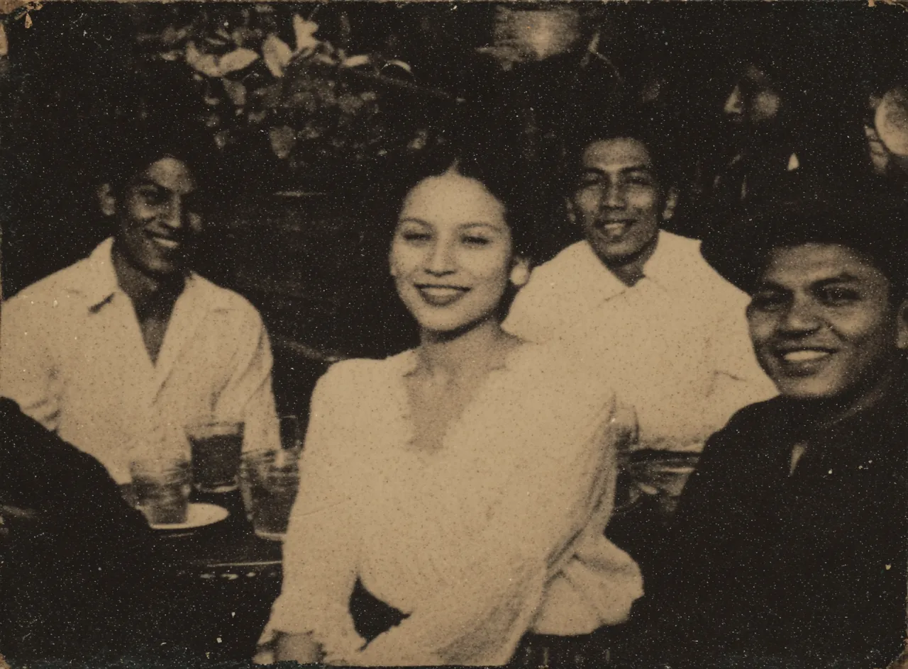 Fotografía en sepia de una mujer sonriente en el centro de un grupo de cuatro hombres sentados a una mesa, con plantas de fondo.