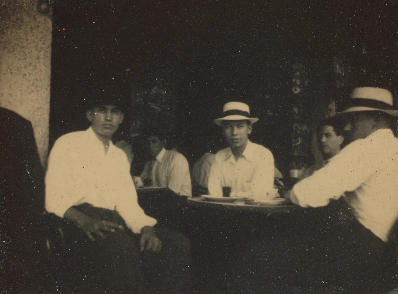 Fotografía en sepia de un grupo de hombres con sombreros sentados alrededor de una mesa con fichas de dominó.