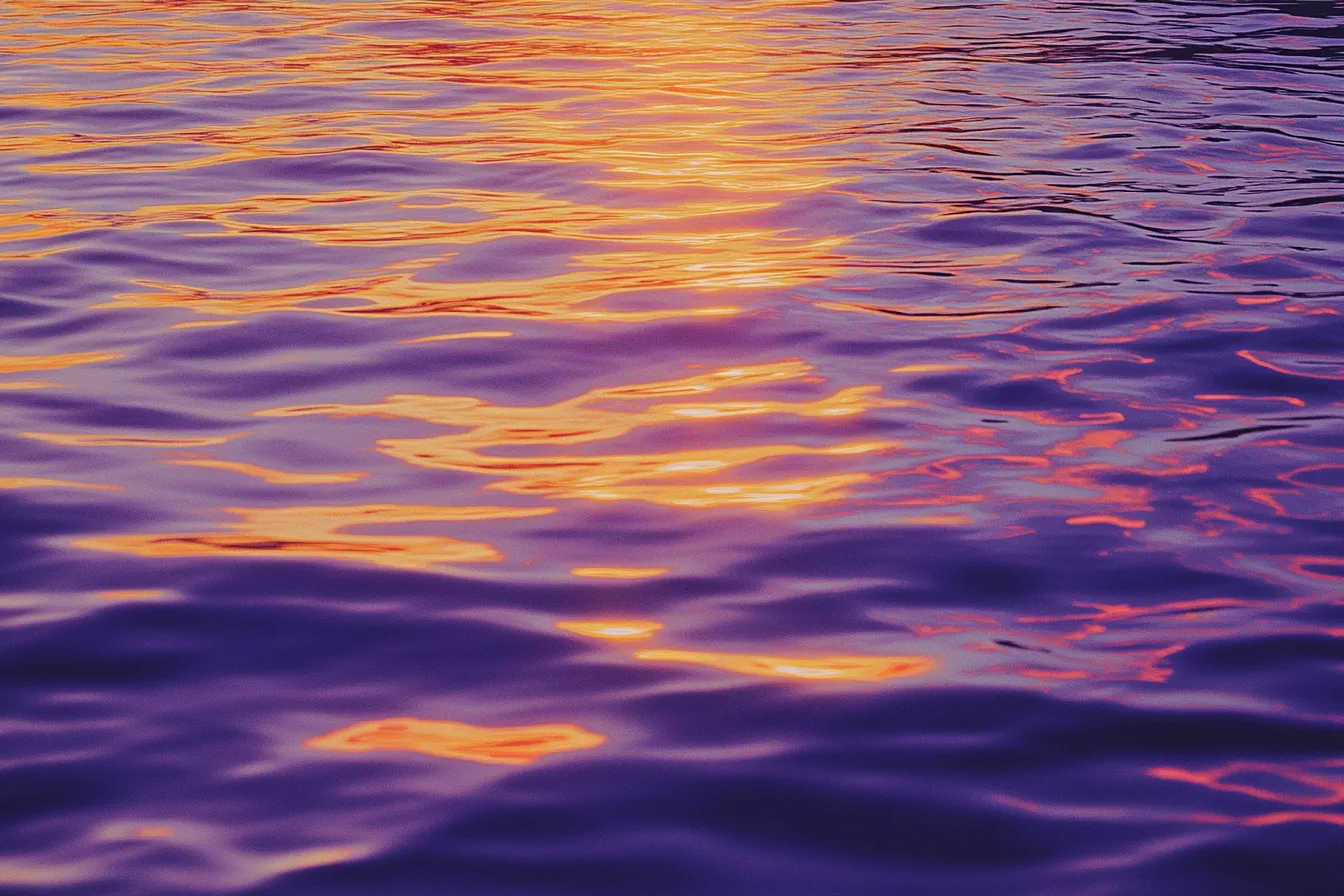 Fotografía de cerca de la superficie del agua con ondas, reflejando luz de atardecer en tonos naranja y amarillo sobre agua de tono púrpura azulado.