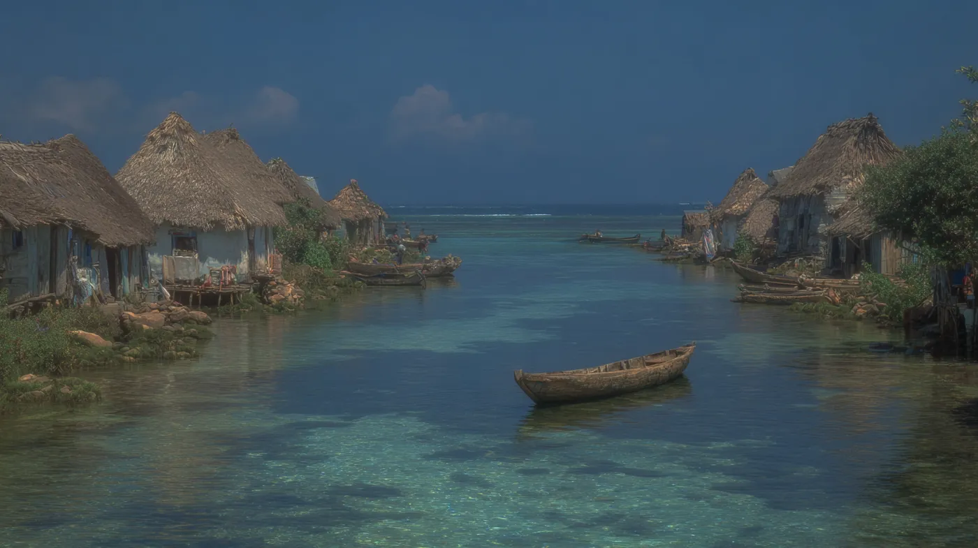 Fotografía de un pueblo de casas de madera sobre pilotes con techos de paja, construido sobre un canal de agua turquesa transparente, con una canoa de madera vacía flotando cerca, bajo cielo azul.