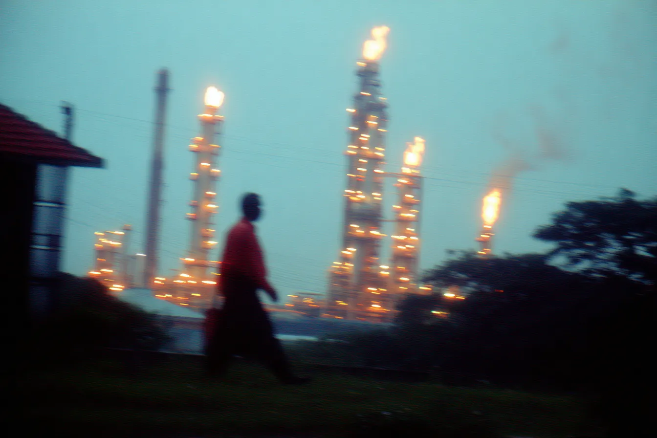 Fotografía borrosa por movimiento de una persona caminando en primer plano vestida de rojo, con una refinería petrolera iluminada y con llamas al fondo, bajo un cielo brumoso al atardecer.