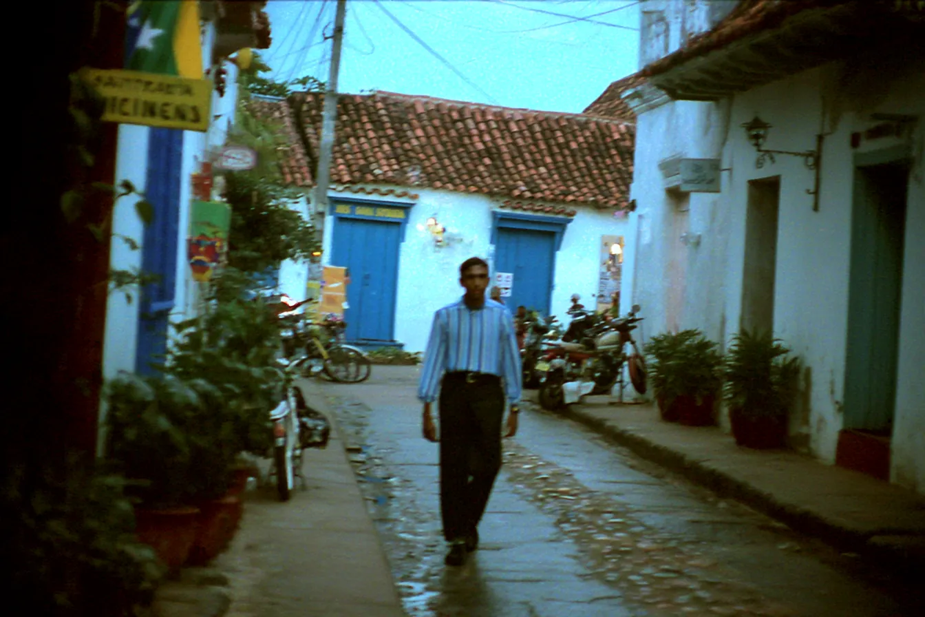 Fotografía de una persona caminando por una calle empedrada de un pueblo colonial, con casas blancas de techos de tejas rojas, puertas azules, motocicletas estacionadas y plantas en macetas, al atardecer.