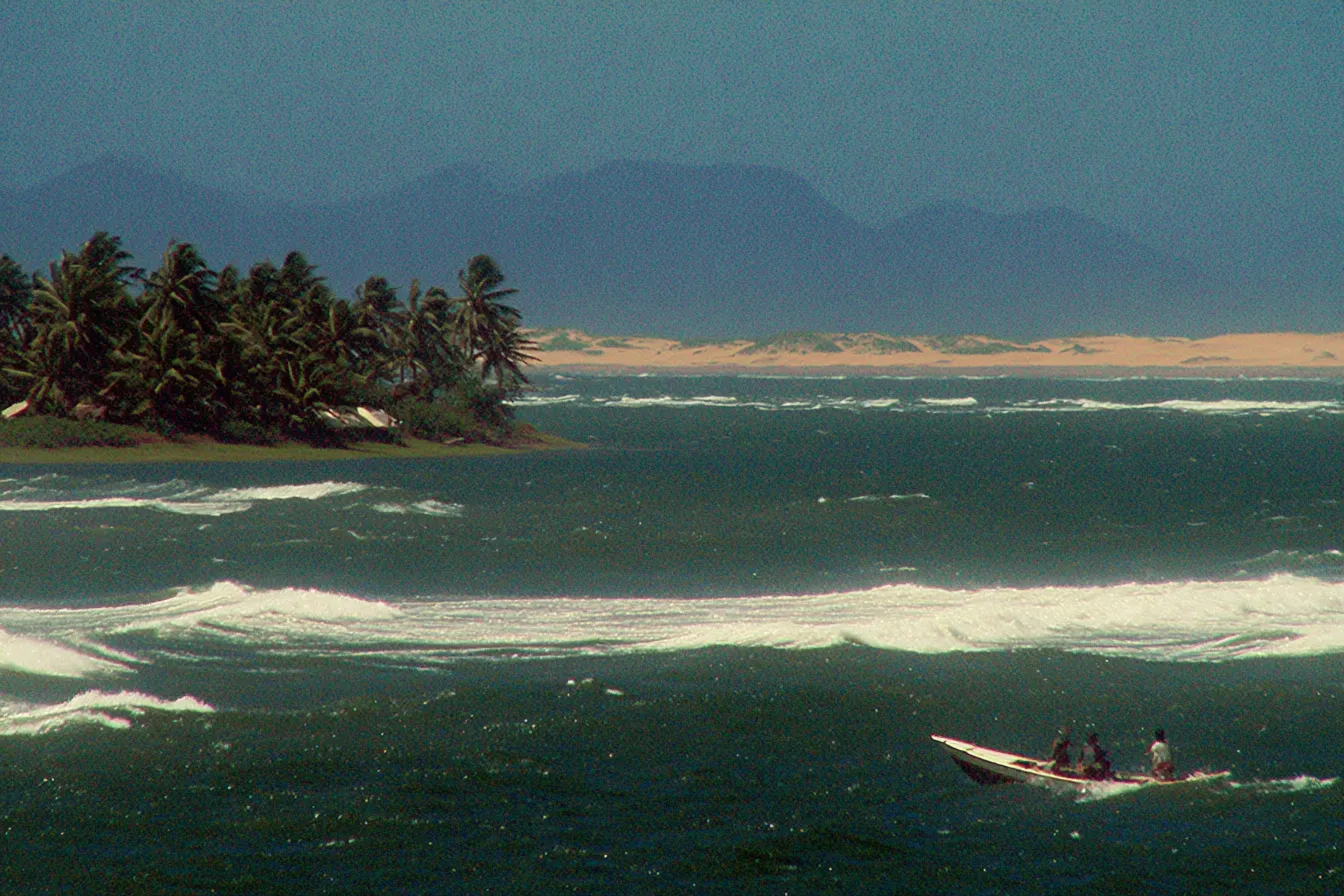 Paisaje costero con una pequeña isla de palmeras, olas rompiendo en la orilla, montañas al fondo y un pequeño bote con dos personas navegando cerca de la playa.