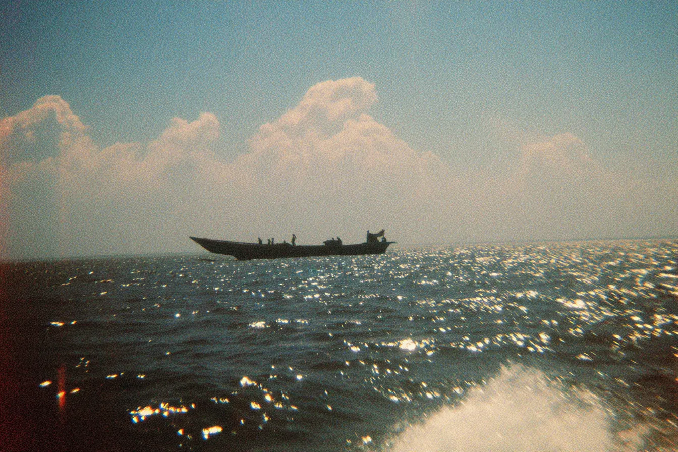 Silueta de un bote de pesca navegando sobre un mar con reflejos brillantes del sol, bajo un cielo con nubes blancas, y espuma de olas en primer plano.