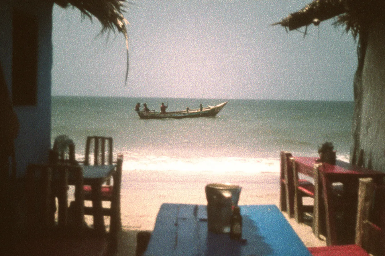 Vista desde una palapa de techo de paja hacia el mar, con un bote de pescadores en el horizonte, sillas de madera y una mesa azul con un vaso en primer plano.