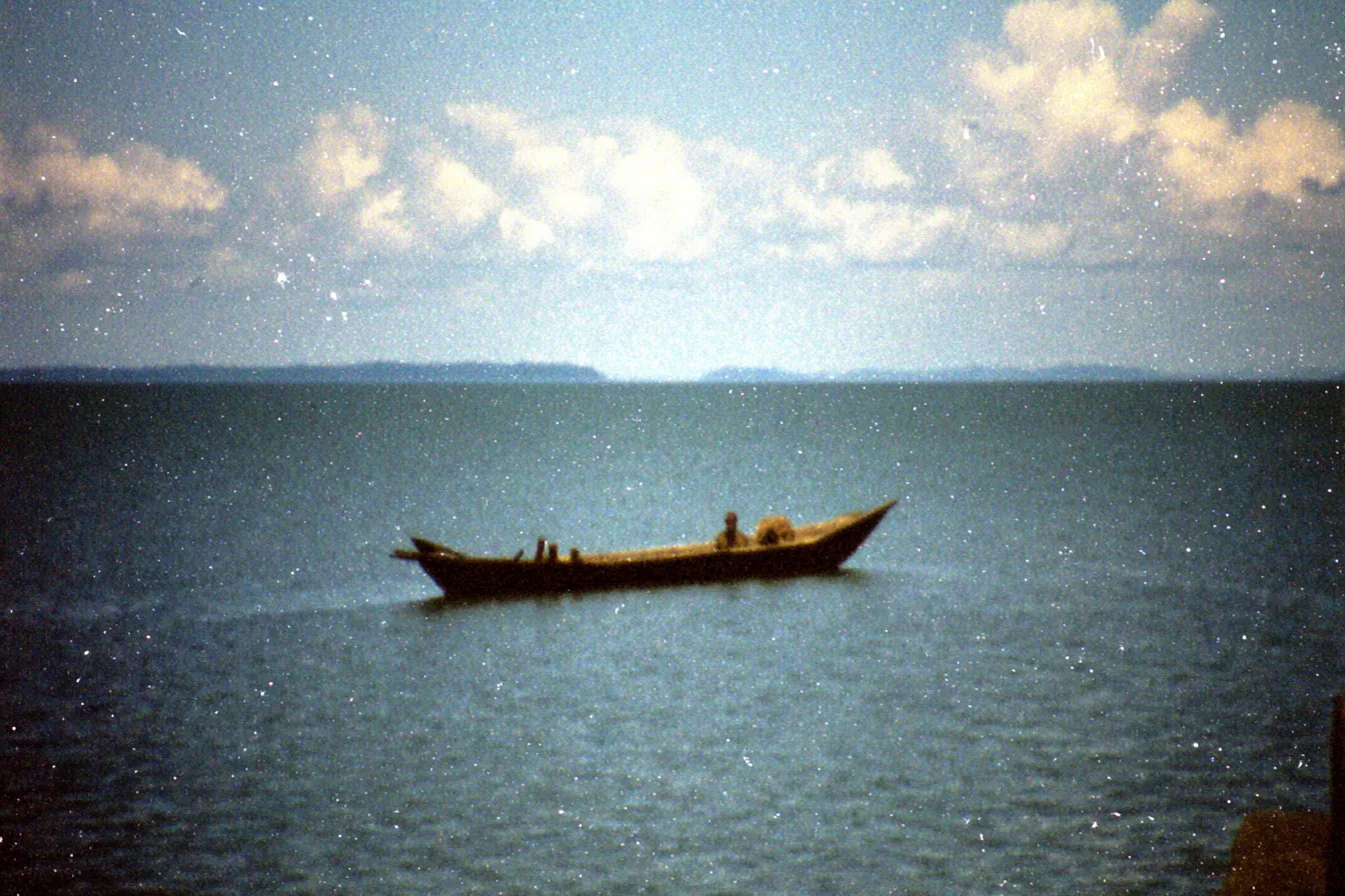Un pequeño bote de pesca navega solo sobre un mar en calma, con una costa lejana y difusa al fondo bajo un cielo nublado.