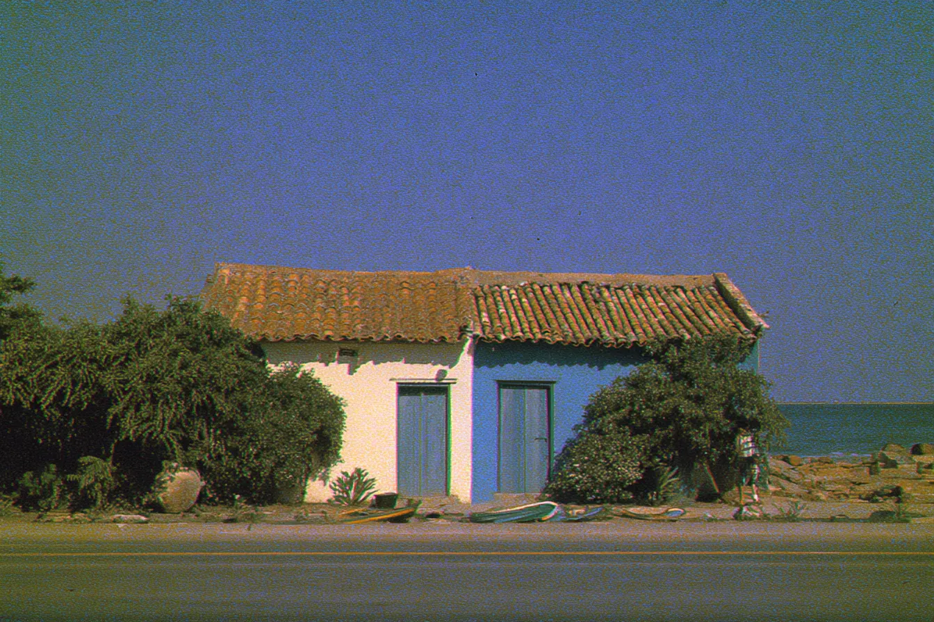 Casa pequeña de paredes blancas y azules con techo de tejas y dos puertas azules, rodeada de arbustos verdes, con una lancha varada cerca y el mar al fondo, bajo un cielo despejado.