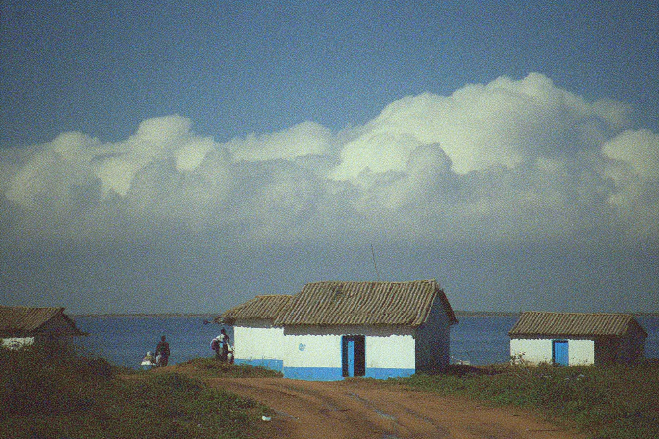 Tres casas con techo de paja y puertas azules junto a un camino de tierra, frente al mar, bajo un cielo con grandes nubes blancas.