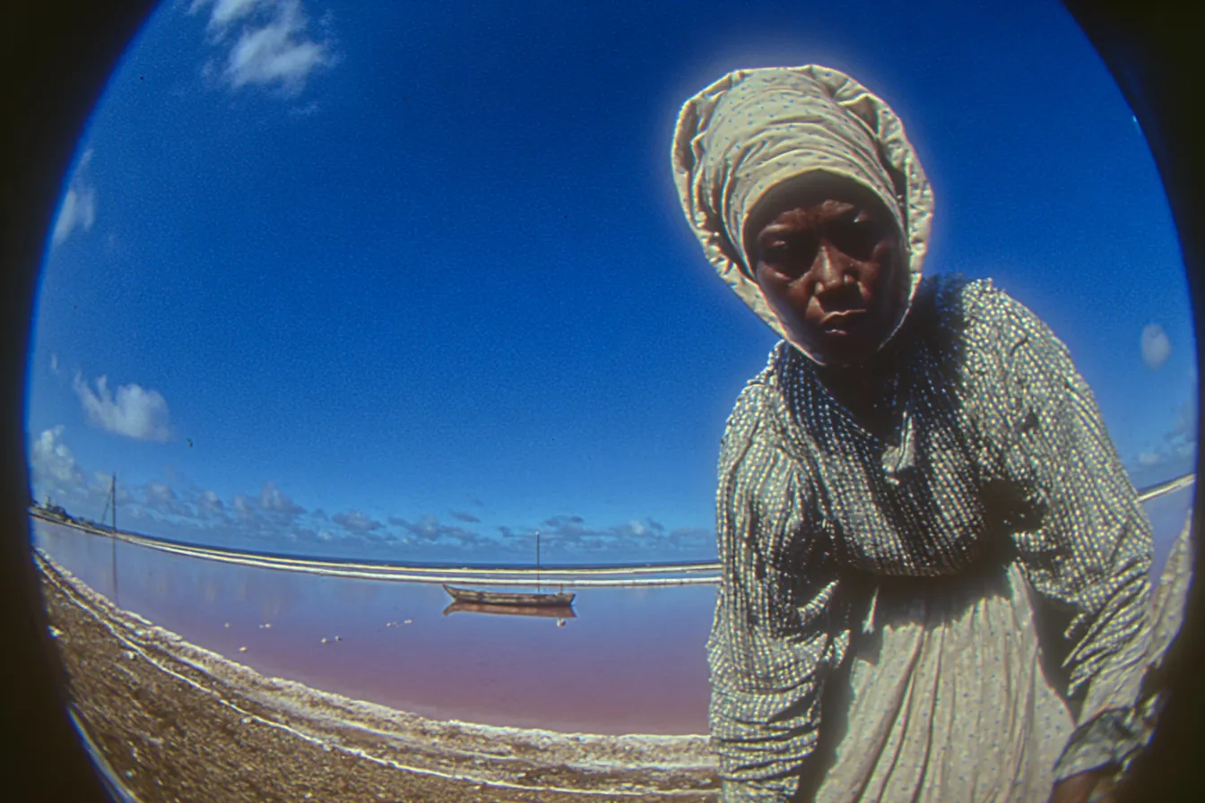 Retrato en gran angular de una mujer con un pañuelo blanco en la cabeza, mirando de frente a la cámara, con un lago de agua rosada y un bote pequeño al fondo, bajo un cielo azul.