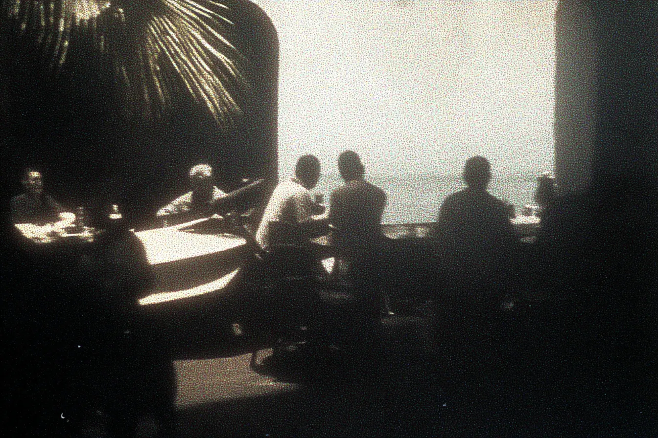 Fotografía en tono sepia de varias personas sentadas en mesas bajo un arco abierto hacia el mar, con una hoja de palmera colgando y una persona tocando un piano.