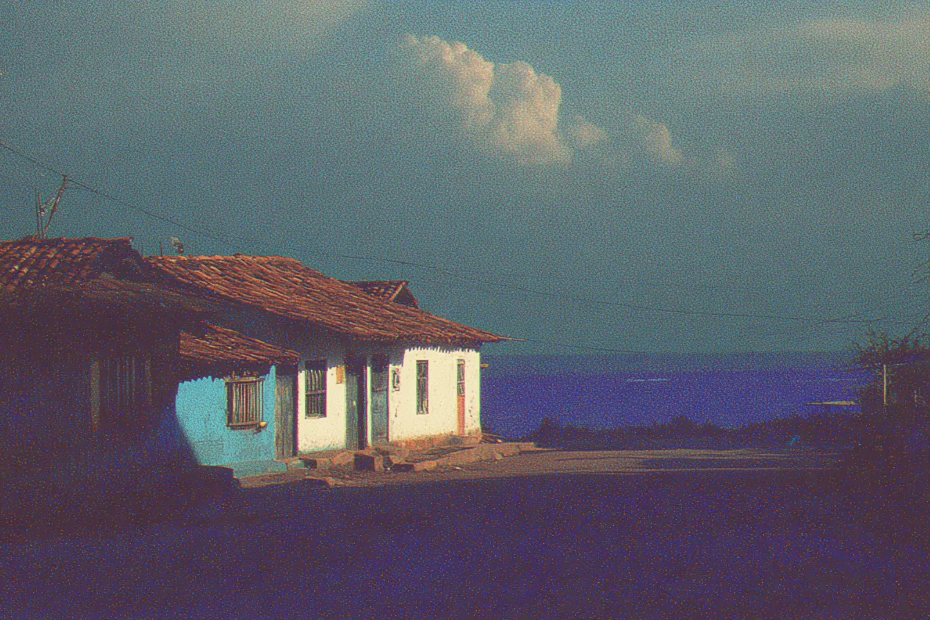 Casa de paredes azules y blancas con techo de tejas, vista al anochecer con un cielo rojizo y morado, el mar visible a lo lejos y cables eléctricos cruzando la escena.