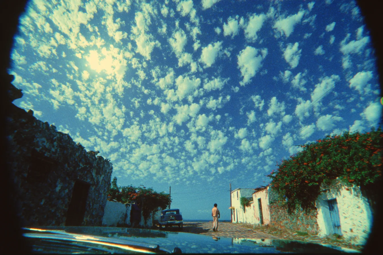 Fotografía en gran angular de una calle de pueblo con casas de piedra, un carro estacionado y una persona caminando, bajo un cielo cubierto de nubes pequeñas y dispersas.