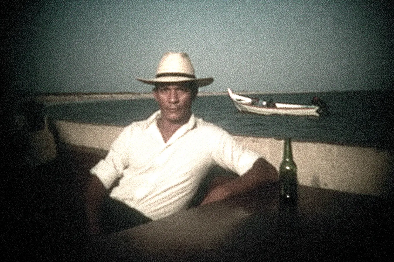 Fotografía antigua de un hombre con sombrero de paja sentado junto a un muro frente al mar, con una cerveza sobre la mesa y un bote de madera flotando detrás.