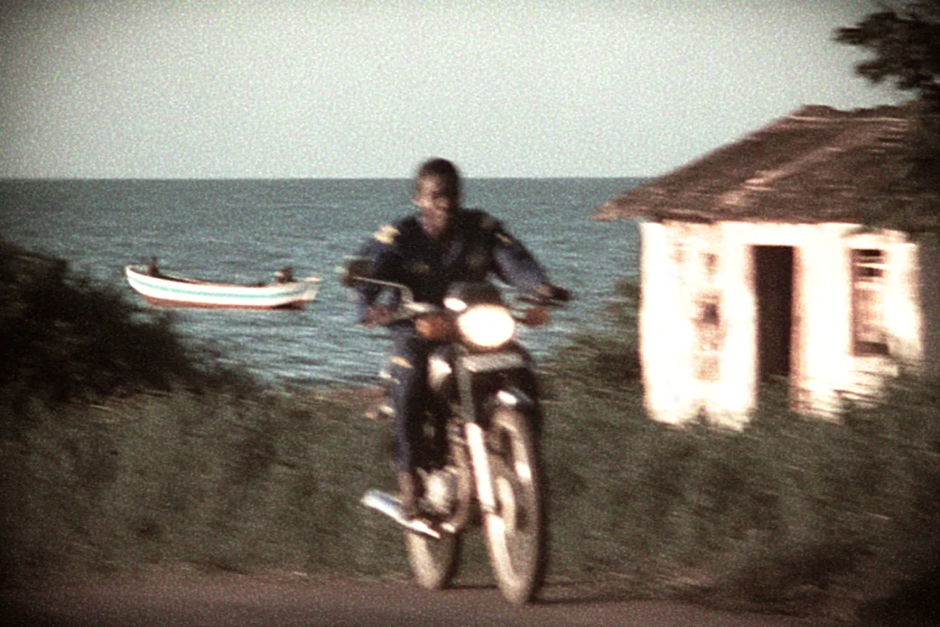 Fotografía antigua de un hombre conduciendo una motocicleta por un camino costero, con una casa pequeña y un bote de madera al fondo junto al mar.