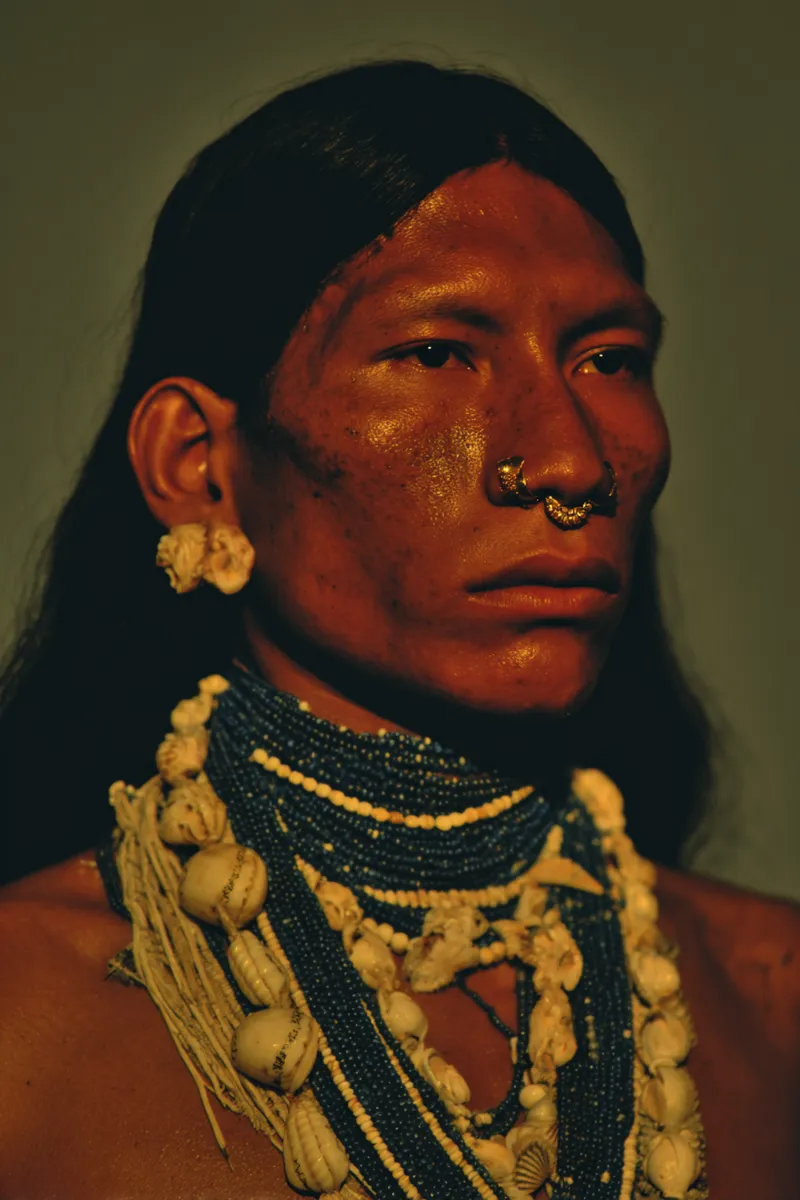 Retrato de un hombre con cabello oscuro, aro dorado en el tabique nasal y collares de cuentas, iluminado de forma tenue contra un fondo oscuro.