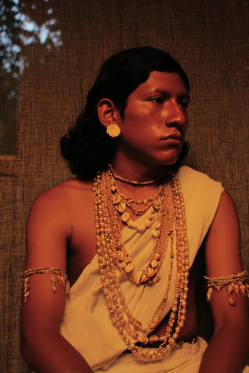 Retrato de un hombre sentado con collares de cuentas superpuestos sobre una túnica tejida color crema, un arete de concha y brazaletes de cuentas en ambos brazos, frente a un fondo de estera tejida.
