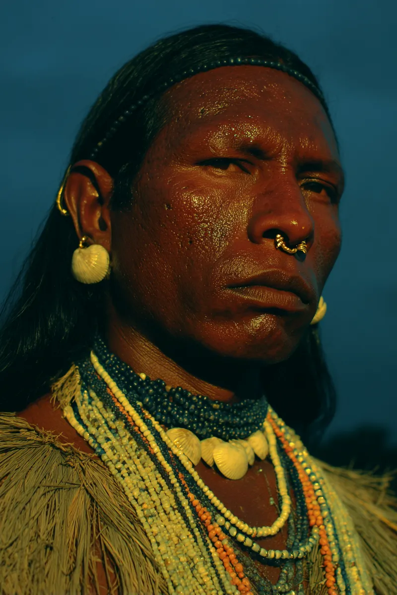 Retrato de un hombre con una vincha de cuentas en la frente, aro dorado en el tabique nasal, un arete de concha y collares de cuentas oscuras y conchas blancas, sobre fondo azul oscuro.