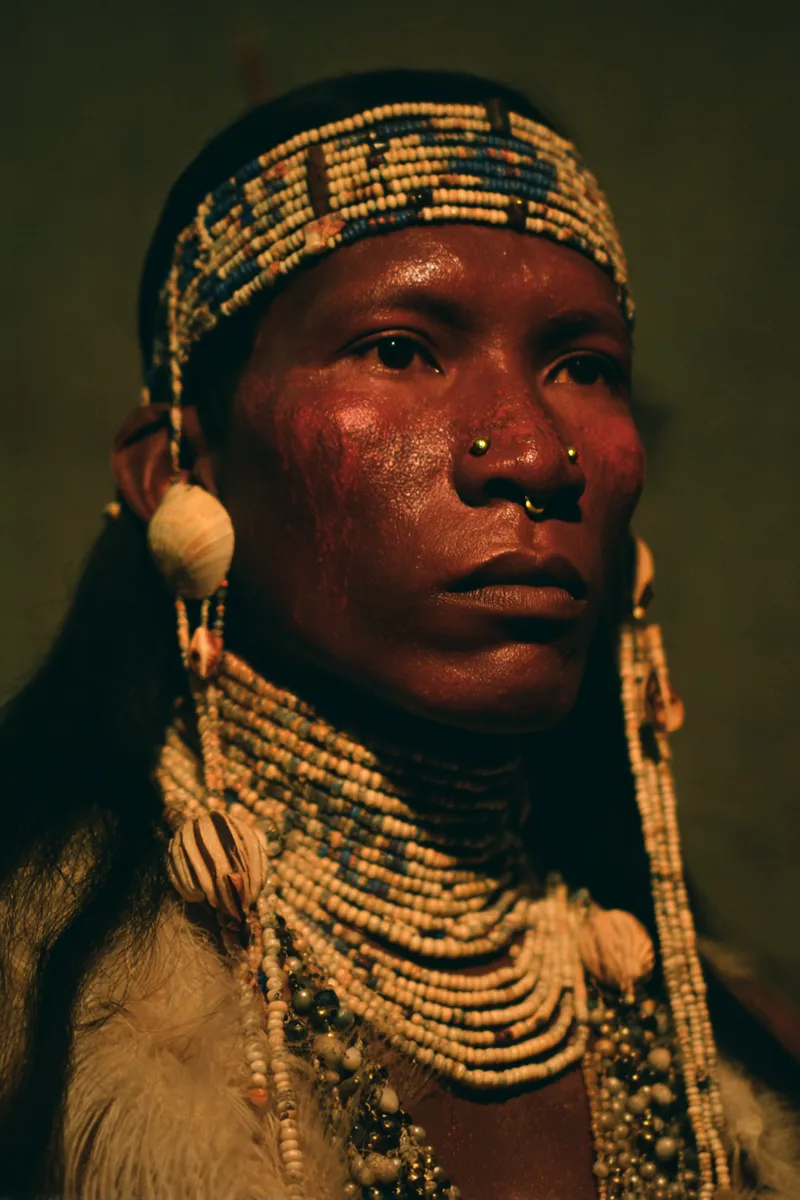 Retrato de un hombre con una vincha de cuentas azules y blancas, piel con tono rojizo, aretes largos de concha y un collar de cuentas, con una prenda de piel sobre los hombros, contra fondo oscuro.