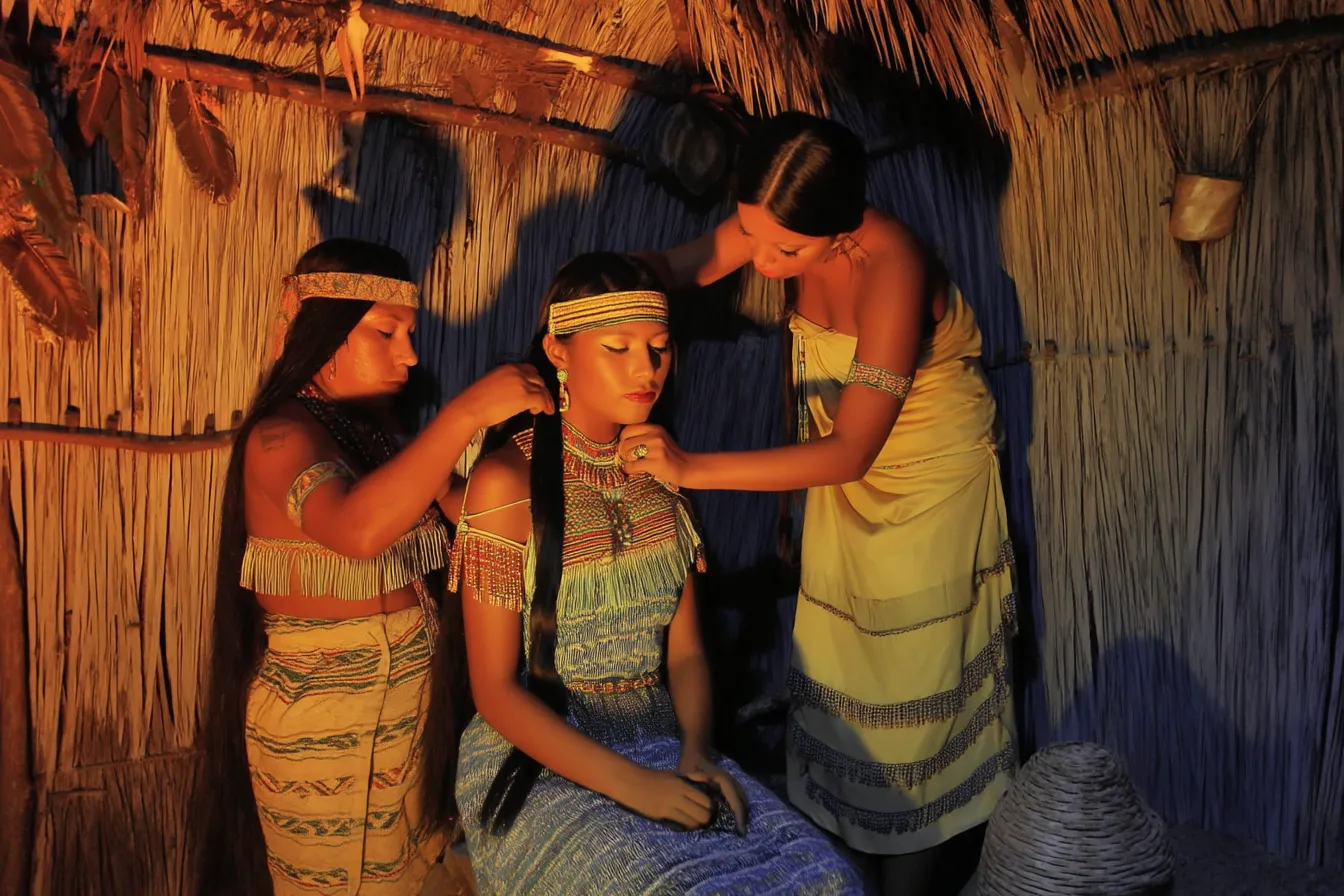 Tres mujeres en un interior de paja: una sentada con top de cuentas y chal a rayas, y otras dos detrás arreglándole el cabello y la ropa con luz cálida y tenue.