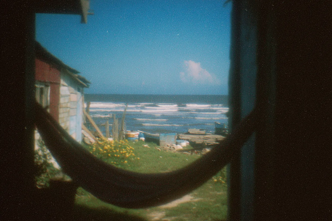 Vista desde el interior de una casa hacia afuera por una puerta, con una hamaca colgada en primer plano y el mar visible al fondo bajo un cielo con nubes, en una fotografía de tono envejecido.