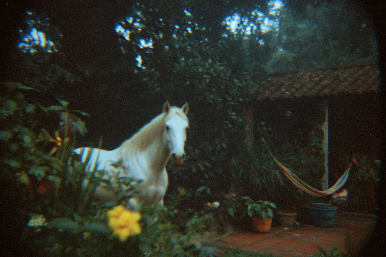 Un caballo blanco de pie entre plantas y flores amarillas en un jardín, con una hamaca y una casa de techo de tejas al fondo, en luz tenue de atardecer y tono de fotografía envejecida.