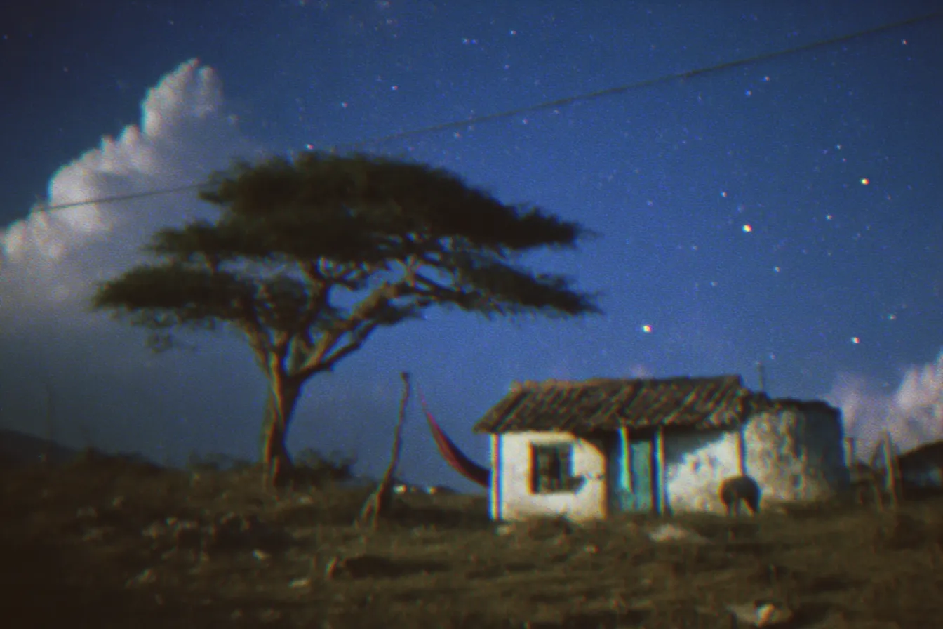 Una casa rural pequeña de paredes claras y techo de tejas junto a un árbol solitario, bajo un cielo nocturno estrellado con nubes, con una hamaca colgada frente a la puerta, en tono de fotografía envejecida.
