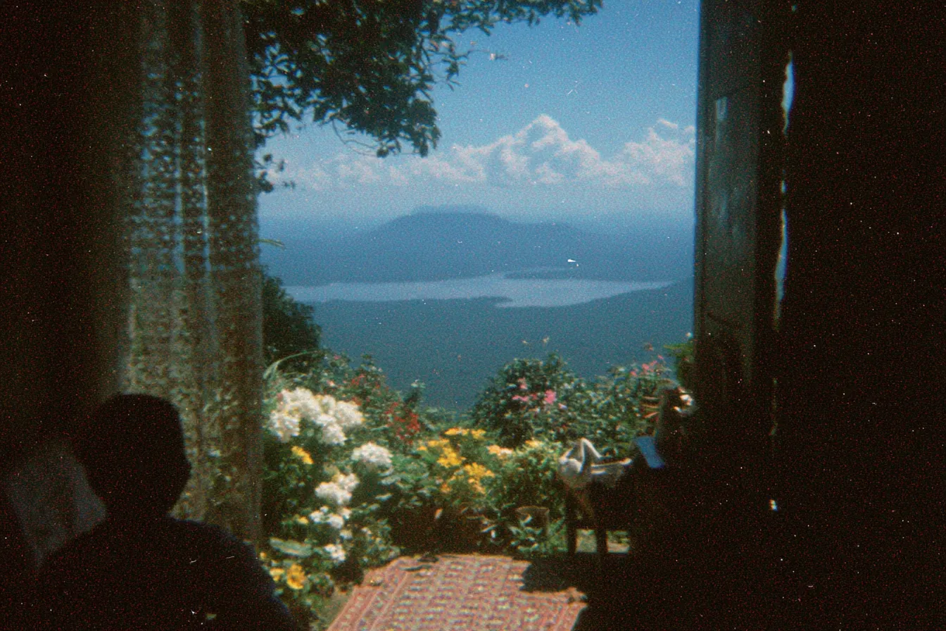 Vista desde un balcón con cortinas hacia un paisaje de colinas, un lago o bahía y cielo con nubes, con flores en primer plano y la silueta de una persona sentada a un lado, en tono de fotografía envejecida.
