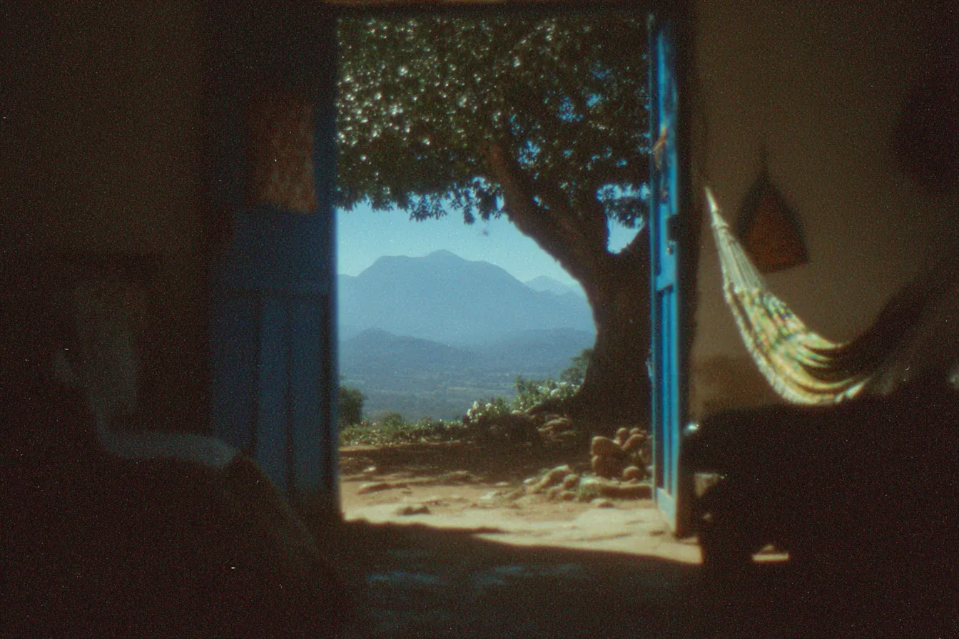 Una puerta azul abierta desde un interior oscuro hacia un paisaje de montañas con un árbol grande, y una hamaca visible a un lado, en tono de fotografía envejecida.