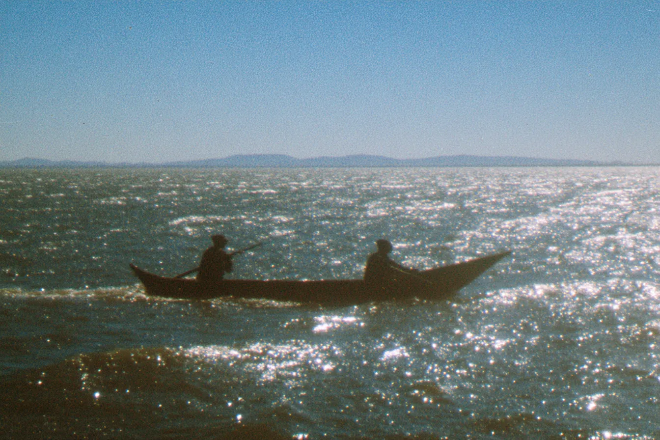 Dos hombres en una canoa de madera, siluetas oscuras sobre un mar que refleja el sol en miles de destellos, con montañas lejanas en el horizonte bajo un cielo despejado.