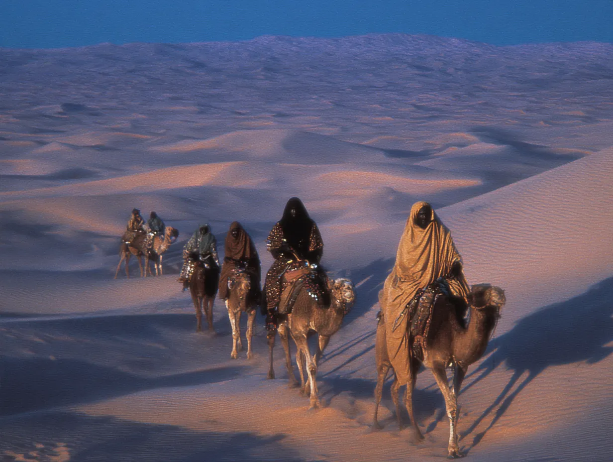 Una caravana de personas montadas en camellos avanza en fila sobre la cresta de una gran duna de arena, proyectando largas sombras al atardecer.