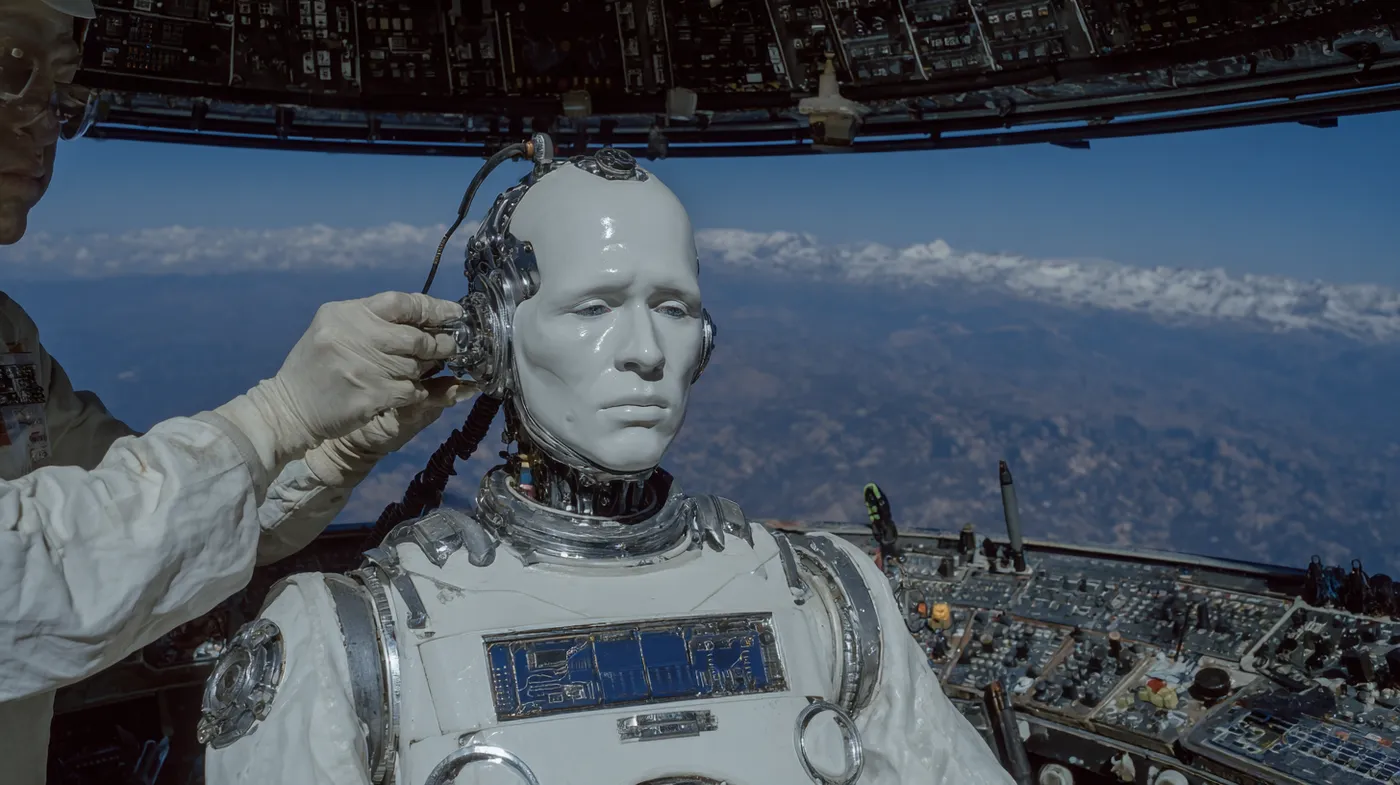 Robot humanoide blanco dentro de un módulo espacial en forma de cúpula, con una mano humana conectándole un cable en la nuca, mientras se ven la Tierra y las nubes a través de las ventanas curvas y paneles de control alrededor.