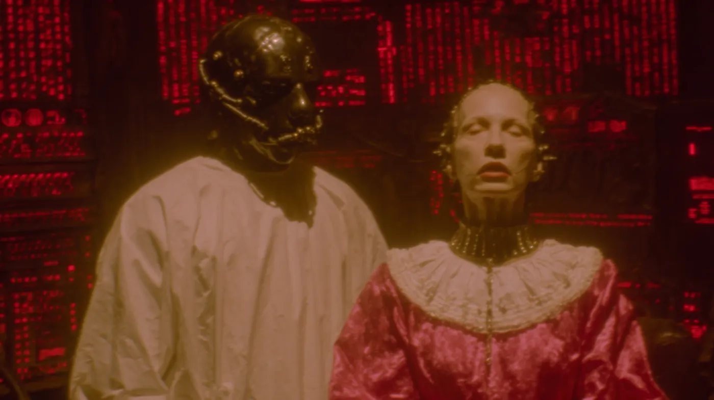 Dos figuras de aspecto robótico con rostros pálidos, una vestida de blanco y otra con un vestido rosado de cuello blanco, de pie frente a paredes cubiertas de texto y números rojos en un entorno oscuro.