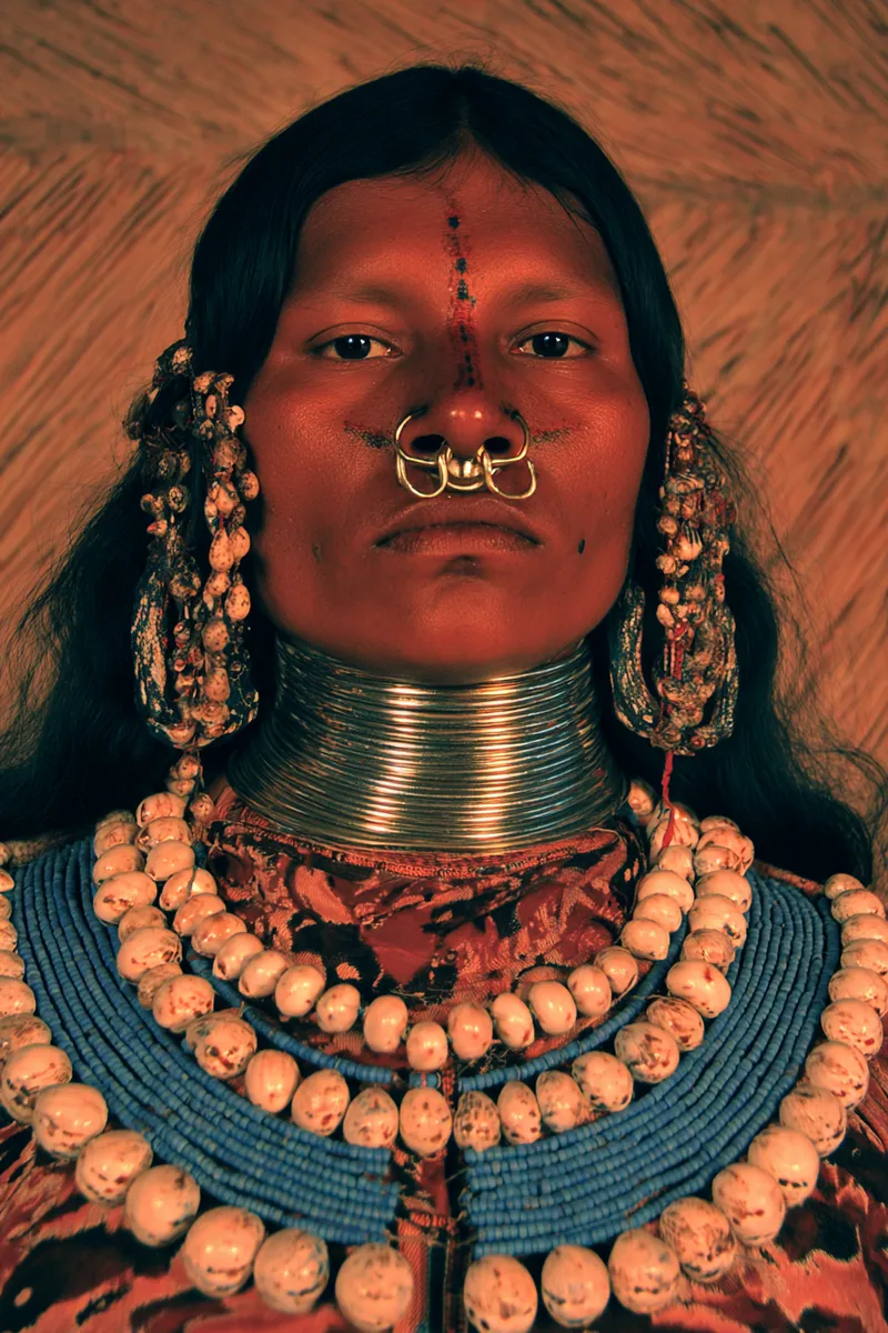 Retrato de una mujer con pintura facial roja en puntos sobre la frente, un aro doble de metal dorado en el tabique nasal, aretes grandes con racimos de conchas, varios anillos metálicos apilados en el cuello y un collar de cuentas blancas y azules, frente a una pared de paja.