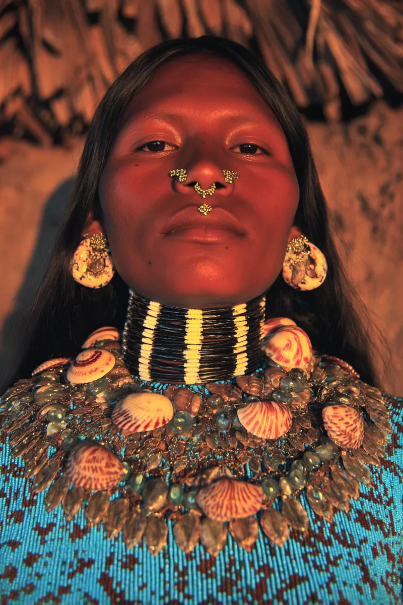 Retrato de una mujer con un aro dorado en el tabique nasal, aretes circulares de concha, un collar de anillos metálicos negros y dorados en el cuello y un collar más grueso de conchas y cuentas sobre un top azul estampado.
