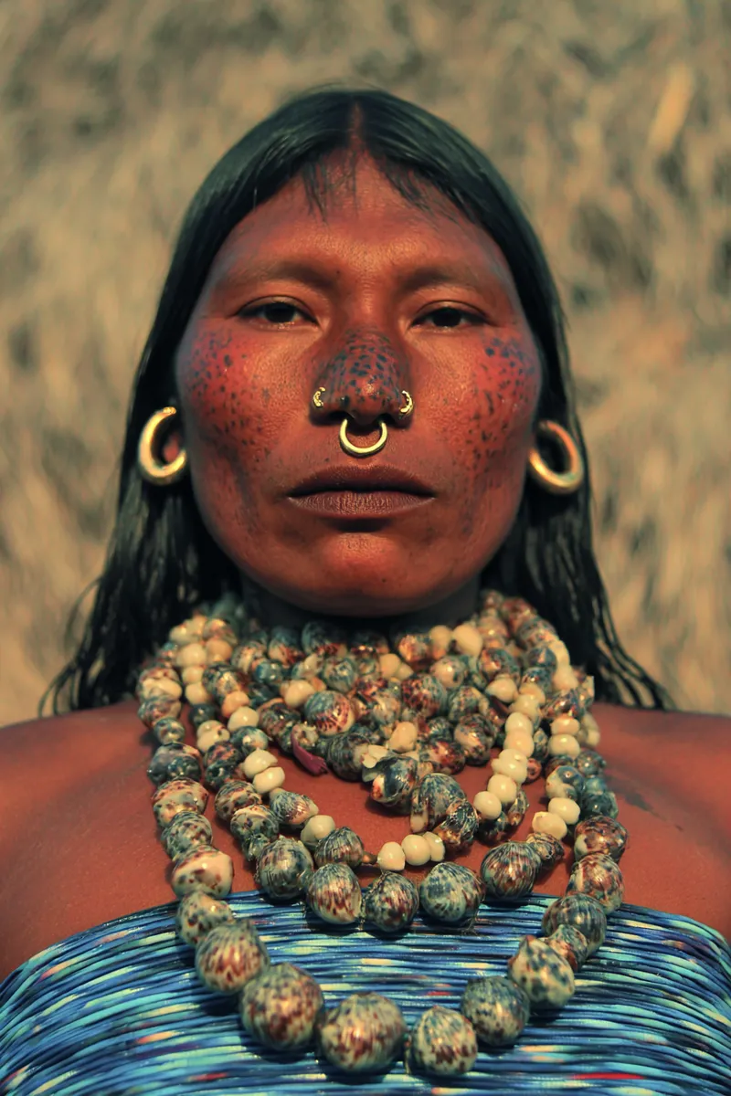 Retrato de una mujer con puntos de pintura roja sobre las mejillas y la frente, un aro dorado en el tabique nasal, aretes blancos en forma de argolla y un collar de varias vueltas de cuentas veteadas sobre un top azul a rayas.