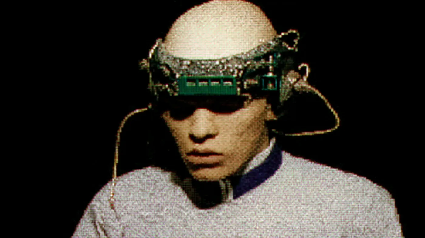 Fotografía en blanco y negro de una persona con la cabeza rapada, que lleva un visor con placa de circuito verde y cables sobre los ojos, vestida con una camisa de cuello azul.