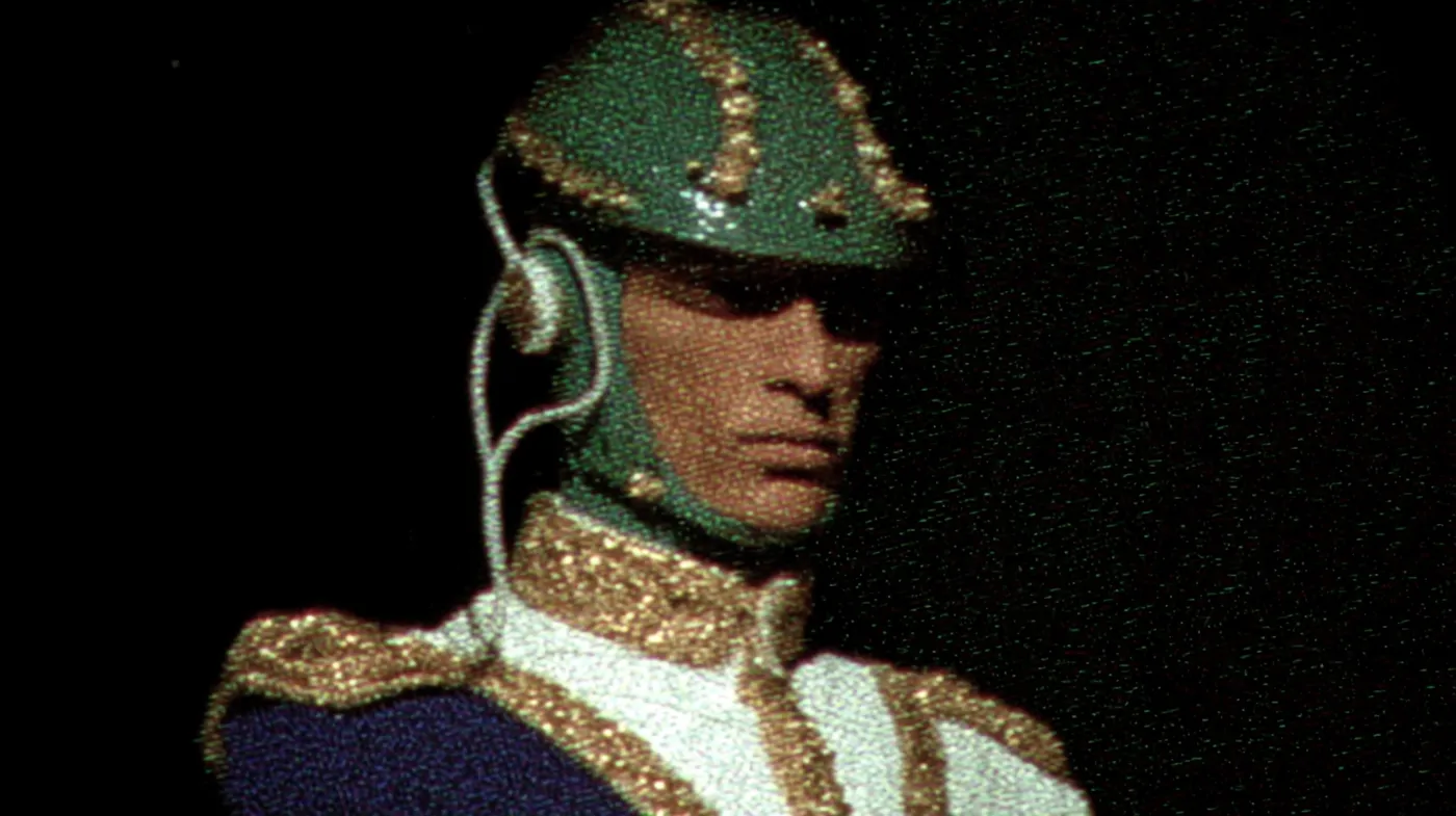 Hombre de perfil con un casco verde cubierto de cuentas y un uniforme blanco con adornos dorados en el hombro, sobre un fondo oscuro.