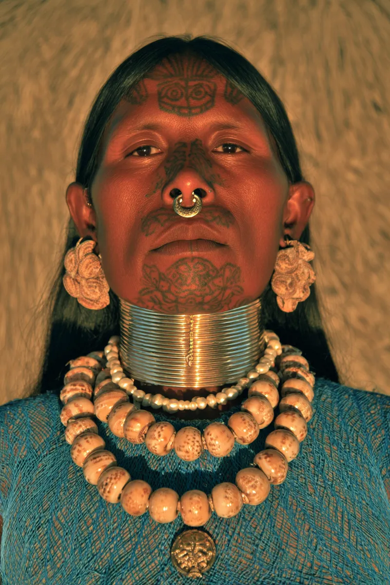 Retrato de una mujer con un símbolo tatuado en la frente y el cuello, un aro dorado en el tabique nasal, aretes con racimos de flores, varios anillos metálicos apilados en el cuello y collares de cuentas sobre un top azul tejido.