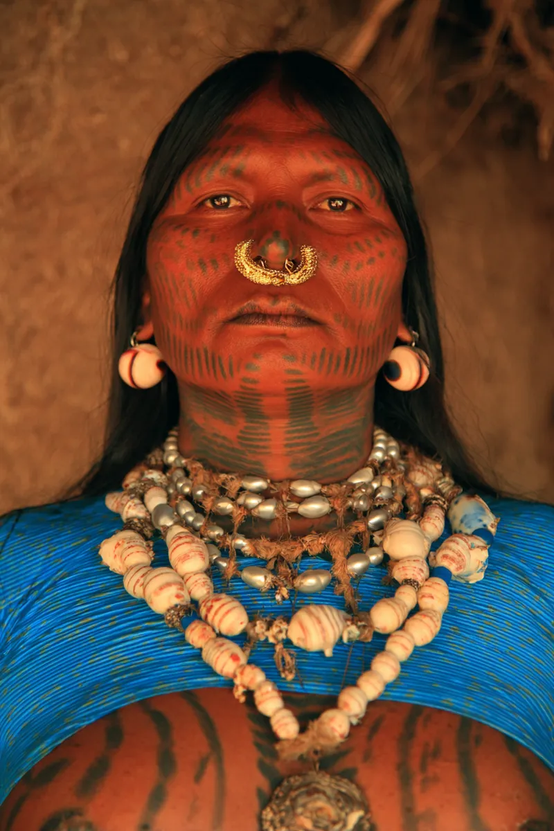 Retrato de una mujer con rayas de pintura facial roja, un adorno dorado con forma de ave en el tabique nasal, aretes redondos con cuentas claras y varios collares de perlas y cuentas superpuestos sobre un top azul texturizado.