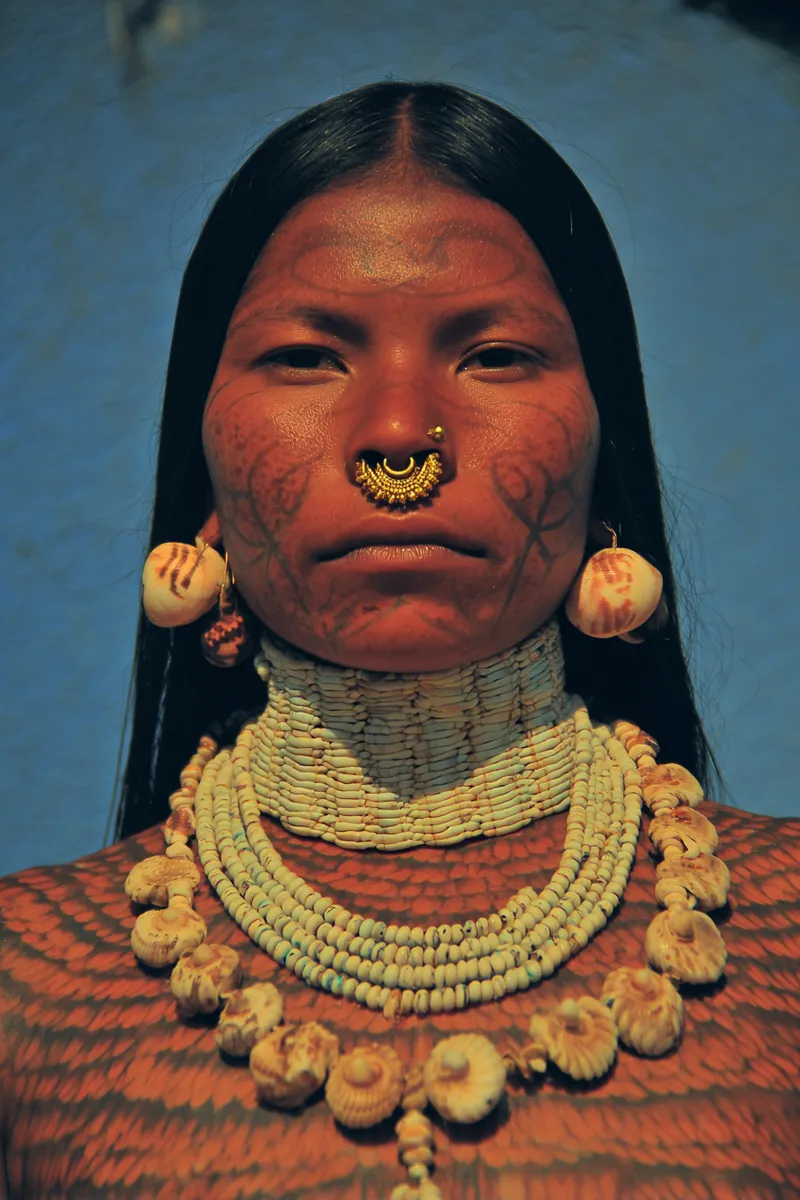 Mujer con pintura facial roja en espiral, arete y anillo de nariz dorados, y un collar grueso de varias vueltas de cuentas de hueso y conchas, sobre fondo azul.
