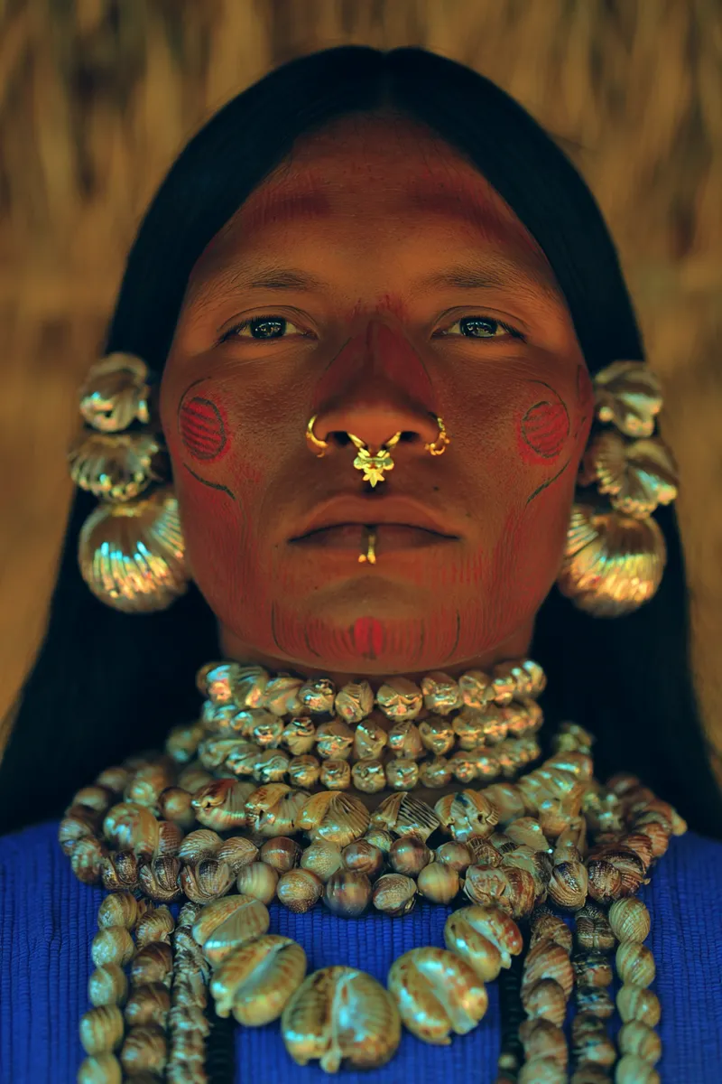 Retrato de una persona con dos círculos rojos pintados en las mejillas, líneas en la frente y la nariz, un arete dorado en forma de flor en el tabique nasal, aretes de concha en espiral y un collar de conchas, con un techo de paja dorado al fondo.