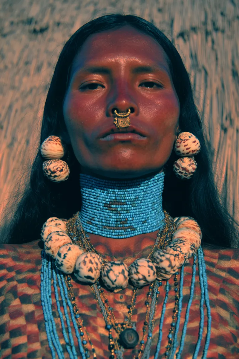 Retrato de una persona con las mejillas pintadas de rojo, un anillo dorado en la nariz, aretes de concha redondos y un collar ajustado de cuentas azules en el cuello, sobre fondo de madera y paja al atardecer.