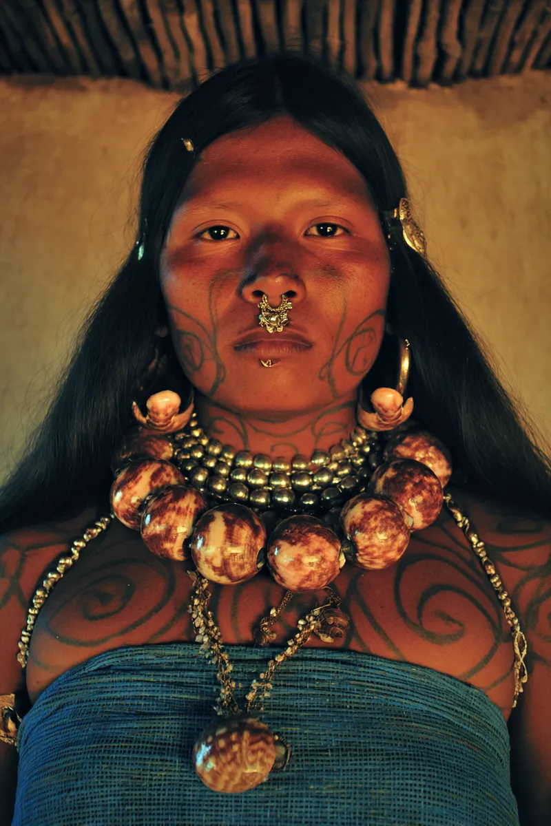 Retrato de una persona con tatuajes en espiral en el rostro, el cuello y el pecho, un aro dorado doble en la nariz, un arete en el labio y un collar grueso de cuentas grandes moteadas, dentro de una choza de techo de paja.