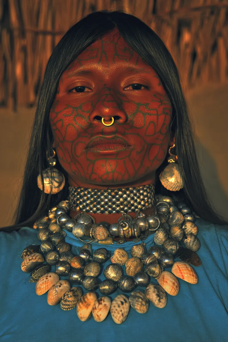 Retrato de una persona con el rostro cubierto por un patrón de pintura facial roja en forma de red, un aro dorado en la nariz, aretes de concha y un collar de cuentas plateadas y conchas, frente a un fondo de paja.