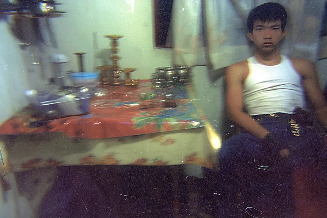 Fotografía granulada de un joven con camiseta blanca sentado junto a una mesa con mantel floreado rojo, ollas y jarras metálicas, en una cocina modesta.
