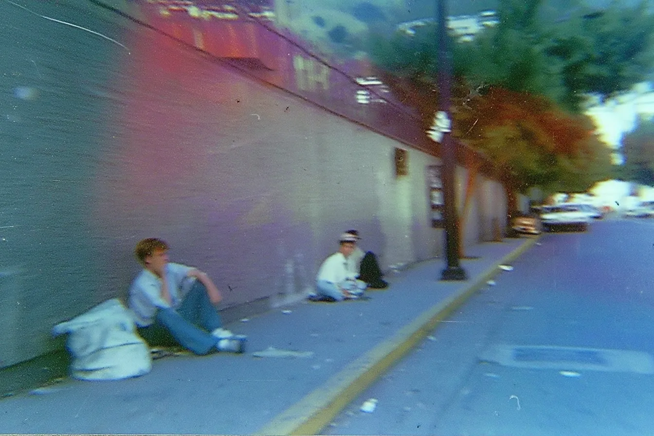 Fotografía borrosa de dos personas sentadas por separado en una acera junto a una pared, con bolsas a su lado, en una calle urbana.