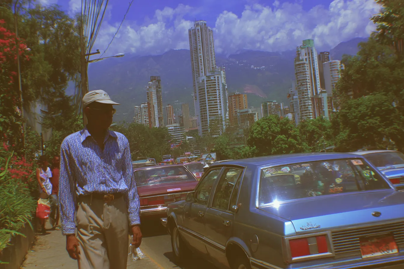 Fotografía de un hombre caminando por una acera con gorra y camisa a cuadros, con edificios altos y una montaña verde al fondo, y autos en tráfico en la calle.