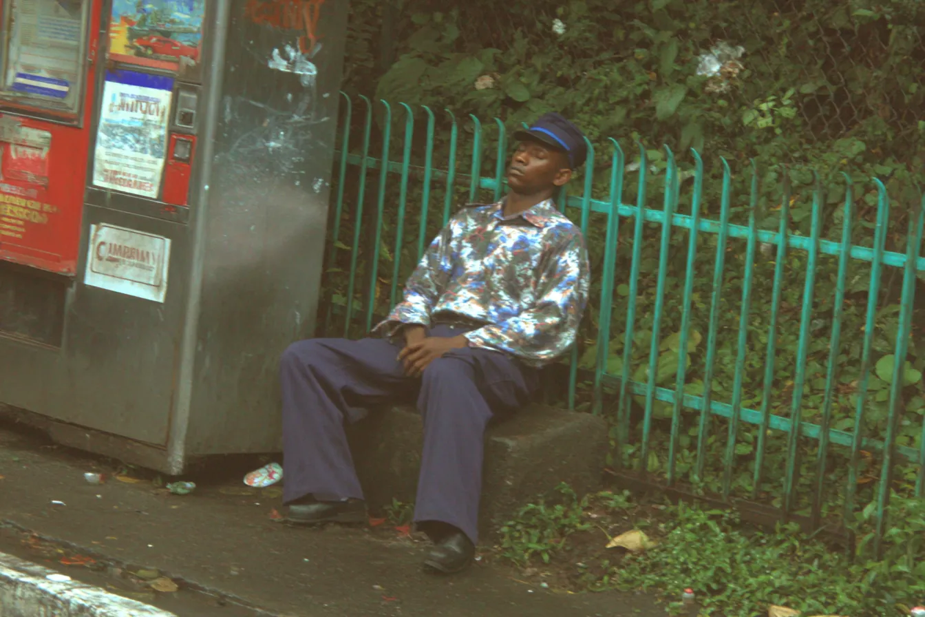 Fotografía de un hombre con gorra y camisa estampada, sentado y con los ojos cerrados junto a una reja verde, cerca de un kiosco de periódicos.