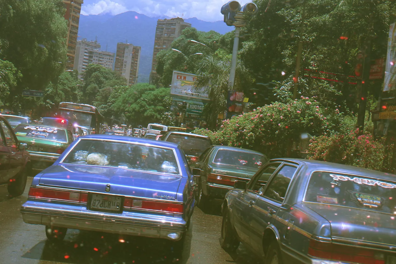 Fotografía de un embotellamiento de autos en una calle de ciudad, con edificios y una montaña al fondo, entre árboles floridos.