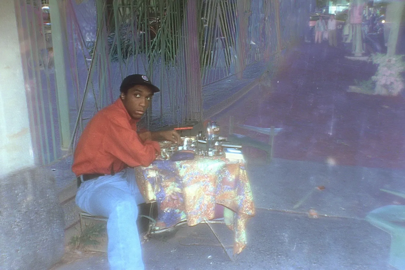 Fotografía de un joven con gorra y camisa roja sentado frente a una mesa cubierta con un mantel de colores, con varios objetos electrónicos a la venta.